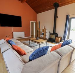 A living room with a beige sectional sofa with blue and orange pillows a wooden coffee table a wall-mounted TV a wood-burning stove and a wooden ceiling at Heritage in St Clears