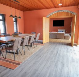 A dining room with a wooden table and eight chairs a rug under the table wooden ceiling and orange walls double doors open to a living room with a sofa and a wall mounted TV at Heritage St Clears