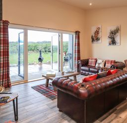 A living room with leather sofas and plaid curtains facing glass doors overlooking a patio and green landscape at Pine Marten in Dornoch