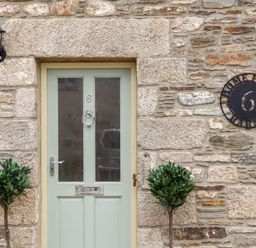 A stone exterior wall with a green double panel door and two small potted trees at Potters Barn in Trevarno near Helston