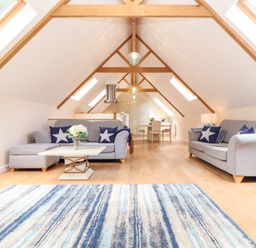 A living area with two grey sofas, a wooden coffee table with flowers, a striped blue rug, and a dining table with chairs at Potters Barn in Trevarno near Helston