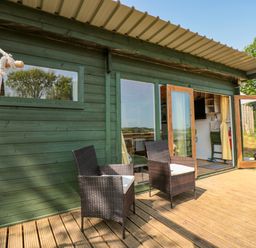 An outdoor wooden deck with two wicker chairs and a hammock outside a green wooden cabin with glass doors open at Dart View Hideout in Totnes