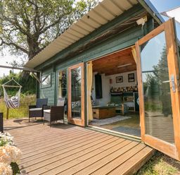An outdoor wooden deck with chairs and a hanging chair next to a small cabin with open wooden framed glass doors at Dart View Hideout in Totnes