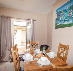A dining room with a wooden table set for four with bowls glasses and a bottle with an open door and grey curtains at The Skipper's House in Weymouth