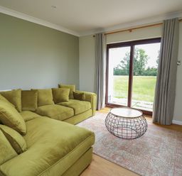 A living room with a green sectional sofa a wireframe coffee table a patterned rug and sliding glass doors with curtains at Sea View in St Clears