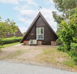 An A-frame house with wooden siding and a gravel driveway surrounded by trees and bushes at Kingfisher in Isleham