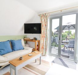 A living room with a cream sofa with blue cushions a wooden coffee table a wall mounted tv floral curtains and glass doors leading to a patio at Kingfisher in Isleham