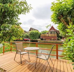 A wooden deck with two chairs and a round table overlooking a water canal with houses and greenery at Kingfisher in Isleham