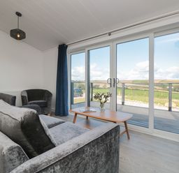 A living room with a gray sofa and chair a wooden coffee table with flowers and a magazine and large glass doors overlooking fields at Bluebell Lodge in Norton near Dartmouth