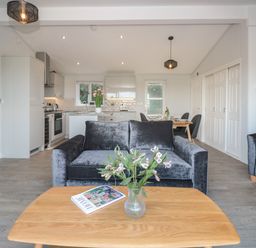 A living room with a gray sofa a wooden coffee table with flowers and magazine and a kitchen and dining area in the background at Bluebell Lodge in Norton near Dartmouth
