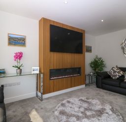 A living room with a mounted television, a wooden panel fireplace, black sofas, a glass console table with a pink flower, and a round rug at Hedd in Amlwch