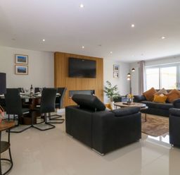A living room with black sofas orange cushions a round wooden table a TV on a wooden panel and a dining table with black chairs at Hedd in Amlwch