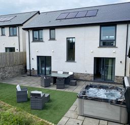 A patio area with outdoor furniture and a hot tub outside a modern white house with solar panels at Galwad y Mor in Broad Haven