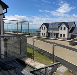 A balcony with two black chairs and glass railing overlooking a street with modern houses and a view of the sea at Galwad y Mor in Broad Haven
