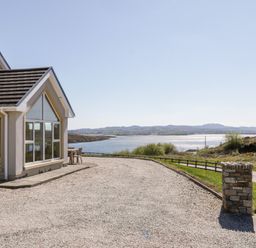 The exterior of a house with large windows next to a gravel driveway and a view of water and hills at Inverbeg Cottage 1 in Downings