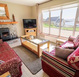 A living room with red patterned sofas a glass coffee table a stone fireplace and large windows overlooking a coastal view at Inverbeg Cottage 1 in Downings