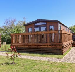 A wooden lodge with a fenced deck and gravel pathway in a grassy area at Mayflower Lodge in Runswick Bay near Staithes