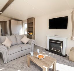 A living room with a gray sofa, wooden coffee table with mugs and croissants, wall-mounted TV above a fireplace and a window at Foxglove Lodge in Runswick Bay near Staithes