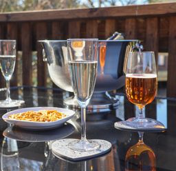 A glass of white wine an amber drink in a glass a bowl of snacks and a metal ice bucket on a glass table on a wooden deck at Tulip Lodge in Runswick Bay near Staithes