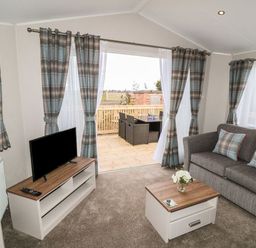 A living room with a gray sofa, plaid cushions, a wooden coffee table, a TV on a stand, and patio doors with plaid curtains at Buttercup Lodge in Runswick Bay near Staithes