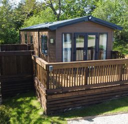 A wooden lodge with a fenced deck surrounded by green trees and grass at Buttercup Lodge in Runswick Bay near Staithes