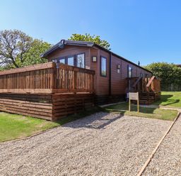 A wooden cabin with a deck and gravel driveway surrounded by grass and trees at Rose Lodge in Runswick Bay near Staithes