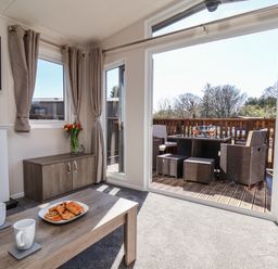 A living room with a coffee table holding pastries and mugs a vase with flowers on a cabinet and outdoor furniture visible through a large glass door at Honeysuckle Lodge in Runswick Bay near Staithes