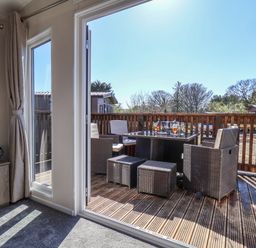 A seating area with a table and chairs on a wooden deck viewed from inside a room with curtains and a cabinet at Lilac Lodge in Runswick Bay near Staithes