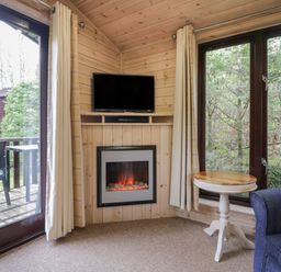 A corner of a room with a wall-mounted TV above an electric fireplace a small round table and a blue armchair near large windows and glass doors at Canadian Cabin 37 in Keswick