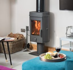 A living room with a wood-burning stove firewood basket and a tray with food and wine on a blue sofa at Shepherds View Dufton near Appleby-in-Westmorland
