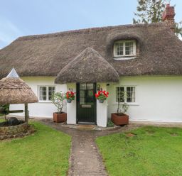 A thatched roof cottage with a front door, two windows, flower pots, a brick well, and a grass lawn at Thatch Cottage in Buckland Newton
