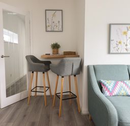 A small dining area with two grey bar stools and a wooden floating shelf table next to a teal armchair with a colorful cushion at The Old Coach House in Conwy