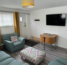 A living room with blue armchairs and sofa a patterned ottoman wooden dining table with chairs yellow curtains and a wall-mounted TV at The Old Coach House in Conwy