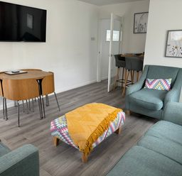 A living room with a sofa armchairs a small table with a yellow throw a wall mounted tv and a small dining table with chairs at The Old Coach House in Conwy