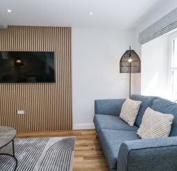 A living room with a blue sofa two cushions a floor lamp a wall-mounted tv and a round table at Riverside Cottage No 3 in Keswick