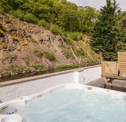 An outdoor hot tub with bubbling water and a wooden picnic table with a bottle and glasses near a hillside at Riverside Cottage No 3 in Keswick