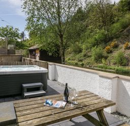 An outdoor patio with a hot tub a wooden picnic table with wine and glasses next to a white wall and trees at Riverside Cottage No 3 in Keswick