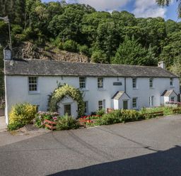 A white long cottage with flower beds and greenery in front surrounded by trees at Riverside Cottage No 5 Keswick
