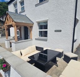 An outdoor patio with cushioned seating and a black glass table in front of a white house with a wooden porch at Briery House in Keswick
