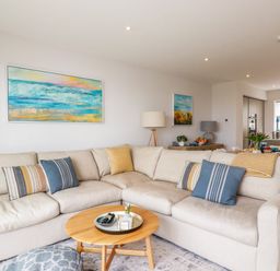 A living room with a beige sectional sofa and pillows and paintings on the wall at 11 Ebbtide in Porth