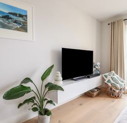 A corner of a living room with a potted plant a wall-mounted TV a basket with cushions and a window at 11 Ebbtide in Porth