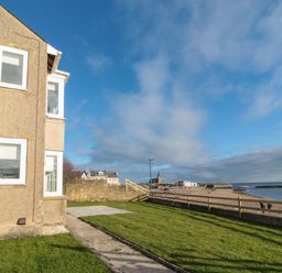 A grassy yard with a concrete path next to a building overlooking a fenced beach and ocean at Fairwinds in Newbiggin-By-The-Sea