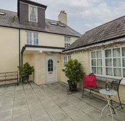 A courtyard with benches two potted plants a small table and a bottle of champagne at Primrose Cottage in Manorbier