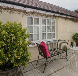 A patio with a black metal bench with pink cushions a small white table with a bottle and glass and two potted plants at Primrose Cottage in Manorbier