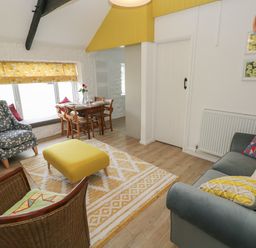 A sitting area with a gray sofa, patterned armchairs, a yellow ottoman, a wooden dining table near a window with yellow curtains at Primrose Cottage in Manorbier