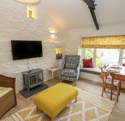 A living room with a yellow ottoman a patterned armchair a wicker chair a dining table set with plates and glasses and a window seat at Primrose Cottage in Manorbier
