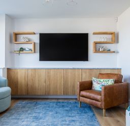A living room with a mounted flat screen tv wood cabinet shelving and armchairs at 12 Tintagel Terrace in Port Isaac