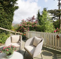 A patio with two wicker chairs a white table with flowers and a wooden fence at Old Farm Cottage in Skelwith Fold