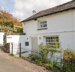 A white cottage with a grey door and small windows surrounded by a low metal fence and stone walls at Old Farm Cottage in Skelwith Fold