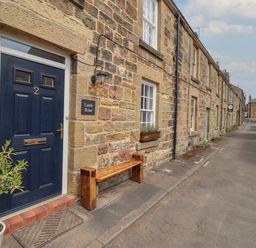A stone row house with a navy door number 2 a wooden bench and a potted plant outside on a street at Lark Rise Alnmouth